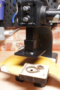 Leather cutting die and yellow vegetable-tanned leather beneath an arbor press foot adapter.
